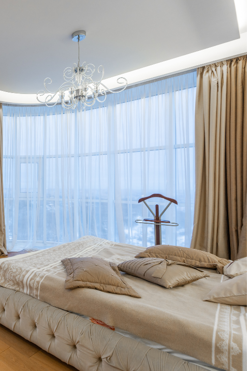 A bedroom with a tufted bed, a chandelier, and elegant layered curtains, showcasing exquisite home furnishing Bangalore design.
