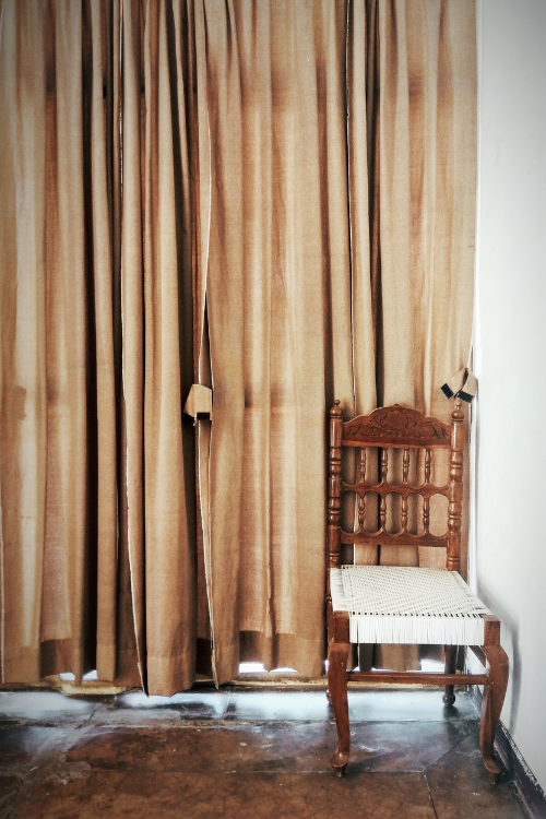 A wooden chair with a woven seat stands in front of heavy, beige curtains, a simple yet charming example of rustic home furnishing.