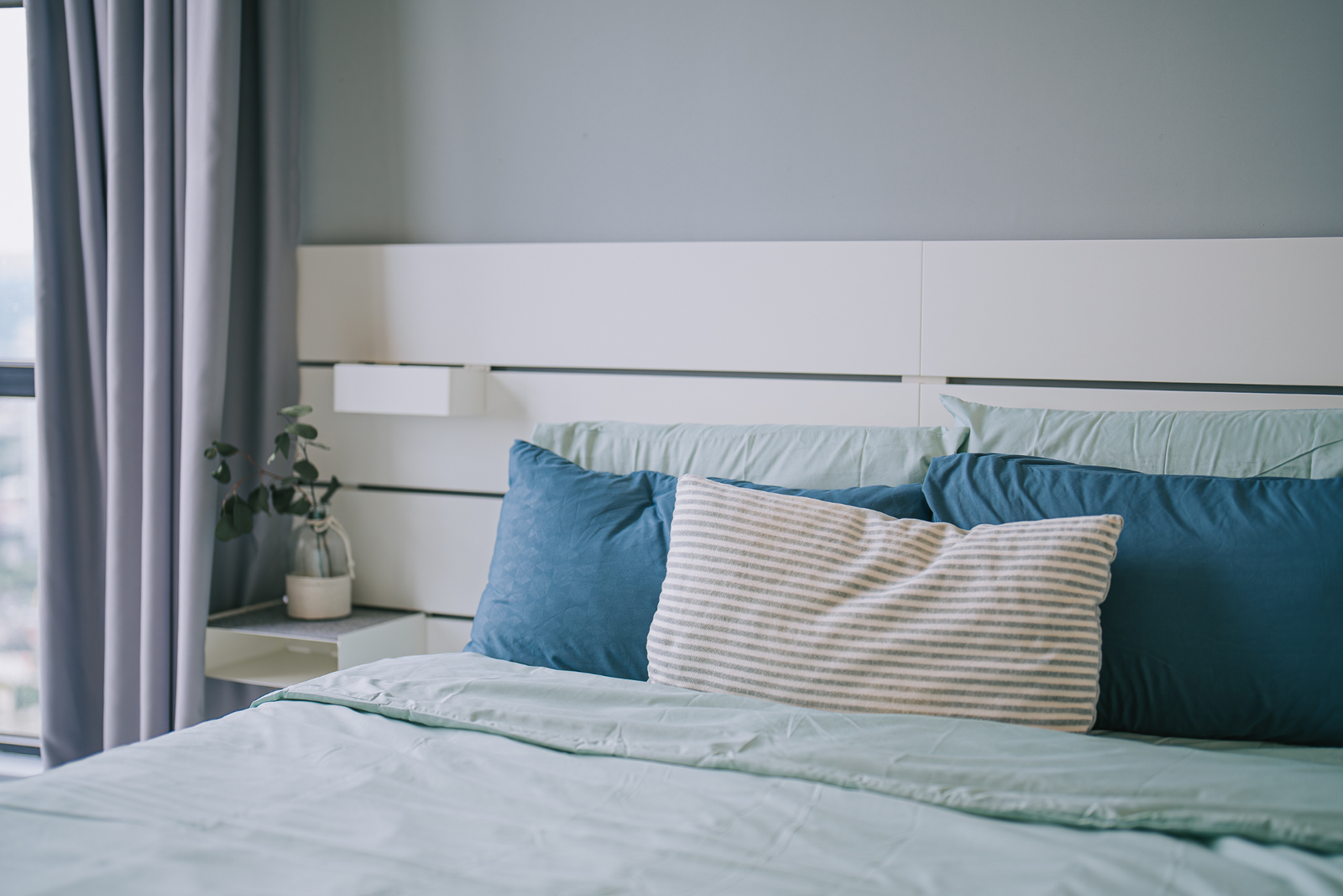 A close-up of a modern bed with light blue bedding, various pillows, and grey curtains, a fine example of home furnishing.