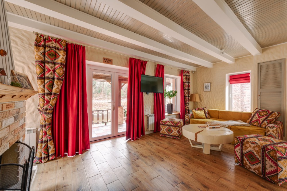 A rustic living room with exposed wood beams, a brick fireplace, and vibrant home furnishing including red and patterned curtains.